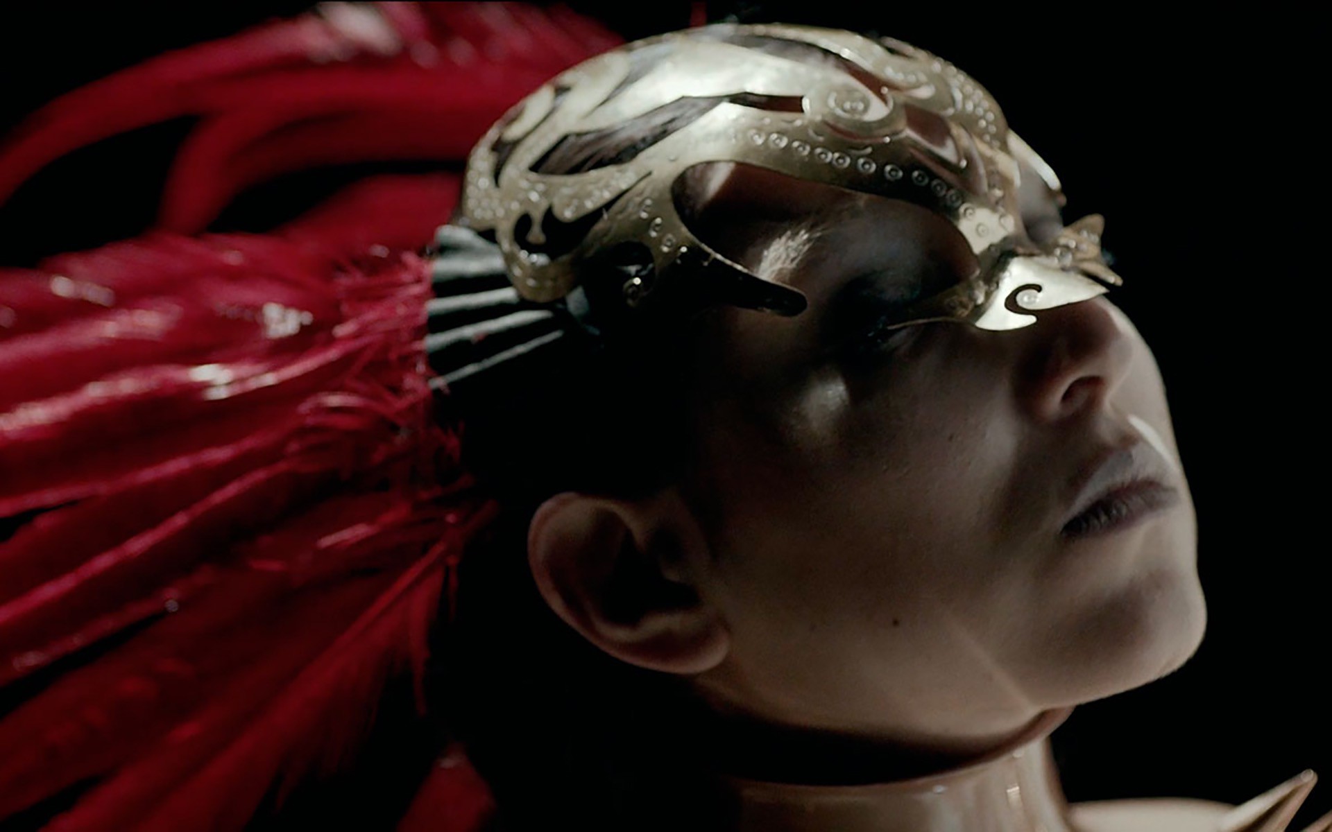 Detalle del fashion film donde se puede ver un primer plano de una ujer con máscara dorada y plumas rojas
